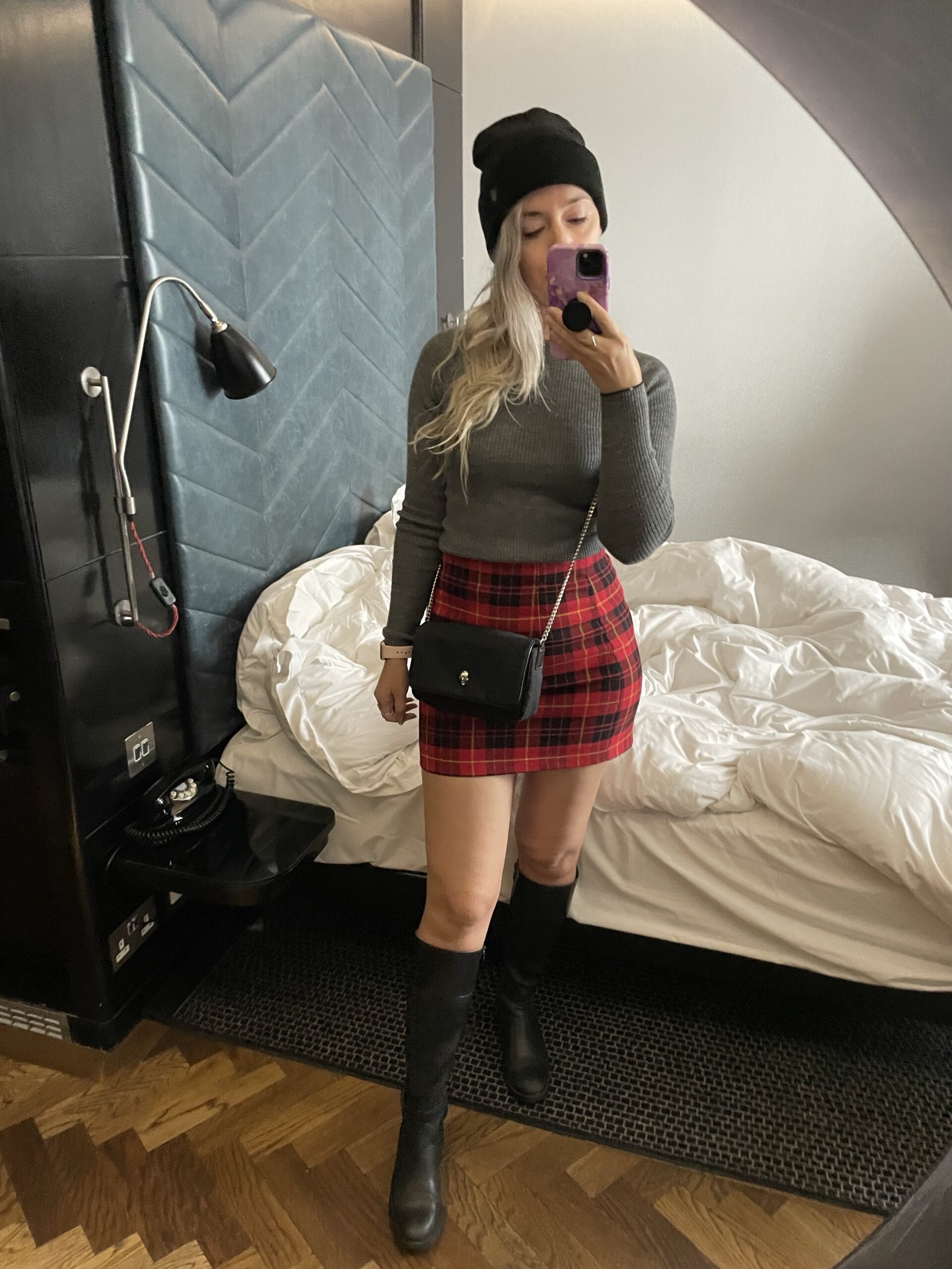 Blonde woman wearing a beanie with a red plaid mini skirt, knee high boots and a grey sweatshirt 